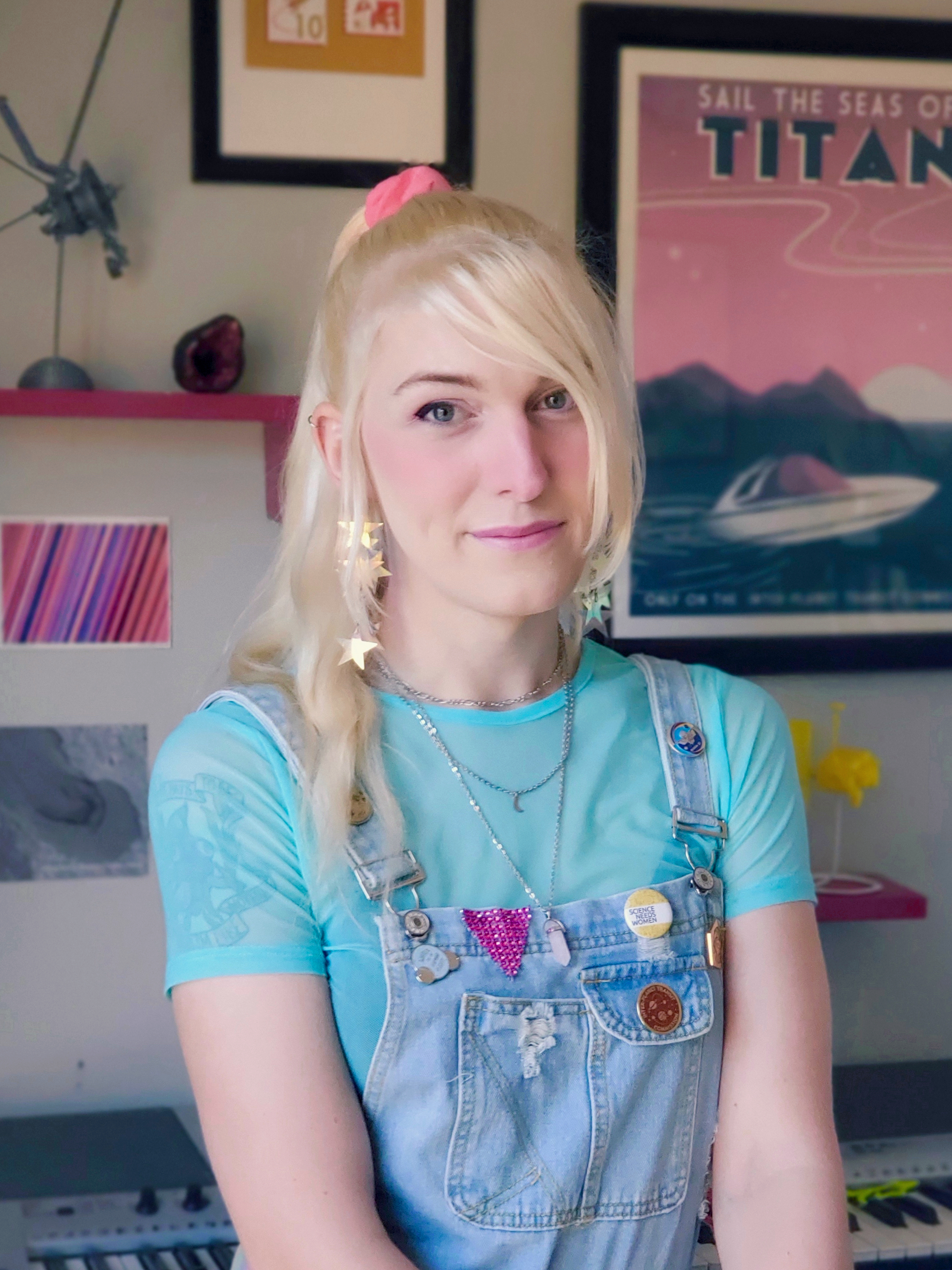 Profile image of Cailin. She has long blonde hair in a ponytail, and is wearing overalls with various space- and science-related pins on them. Behind her are several of her favourite knick-knacks, including rocks, posters, and spacecraft models.